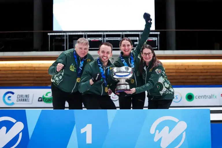 Four Caucasian adults, two male and two female, all wearing Australian team branded jumpers in dark green, stand behind a blue banner with the number 1 on it, holding a large cup trophy. They all smile to camera with their hands in fists or in the air.