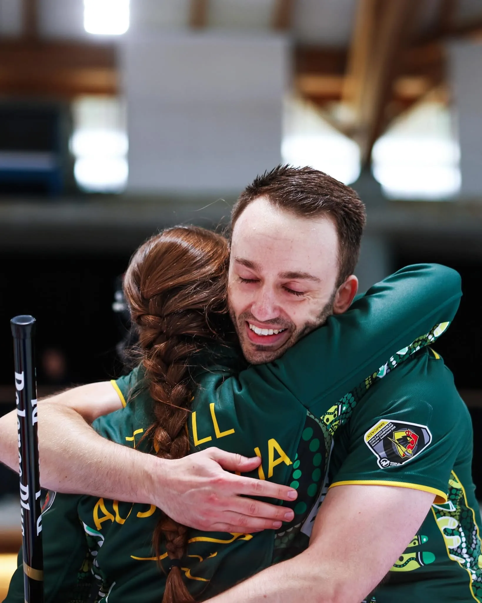 A male athlete faces the camera hugging his female teammate who faces away from the camera. She has a long brown plait in her hair, they both wear green Australian team kit uniform.