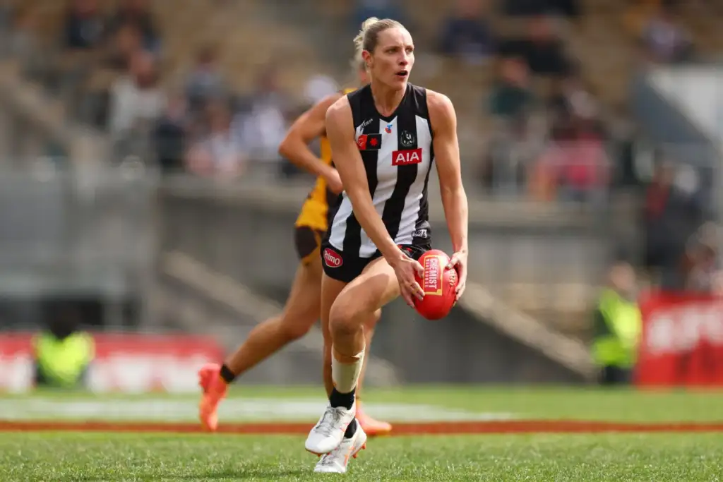 A female athlete with blonde hair tied up is in action playing AFLW. She wears a black and white striped guernsey and black shorts, and is running whilst holding a red football in her hands. A green pitch and grandstand can be seen blurred in the background.