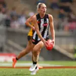 A female athlete with blonde hair tied up is in action playing AFLW. She wears a black and white striped guernsey and black shorts, and is running whilst holding a red football in her hands. A green pitch and grandstand can be seen blurred in the background.