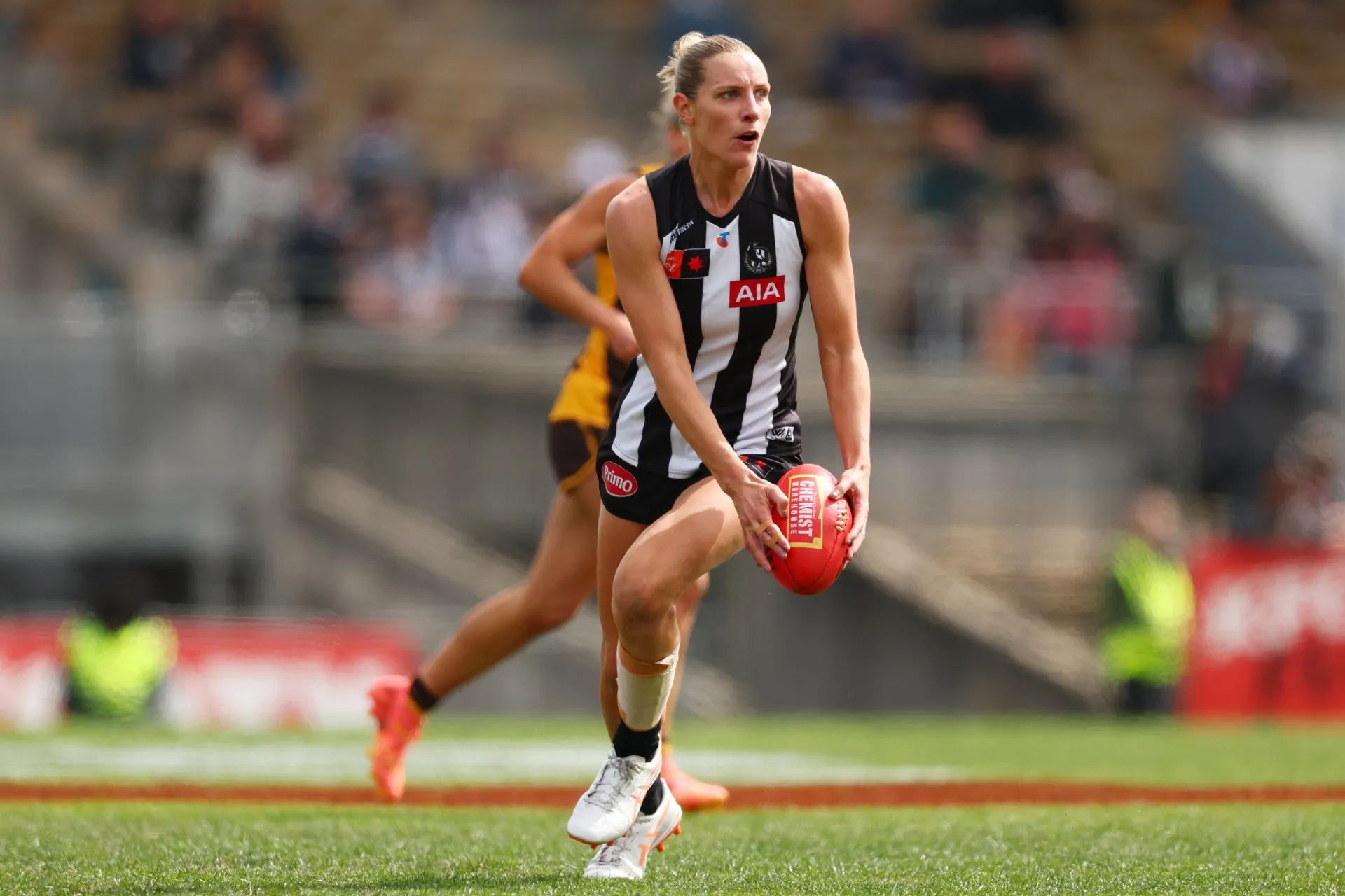 A female athlete with blonde hair tied up is in action playing AFLW. She wears a black and white striped guernsey and black shorts, and is running whilst holding a red football in her hands. A green pitch and grandstand can be seen blurred in the background.