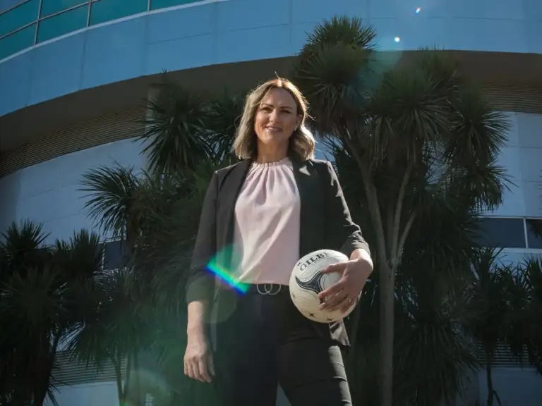 A woman with shoulder length brown hair stands in sunlight wearing a light pink top and navy blazer, holding a netball to her hip.