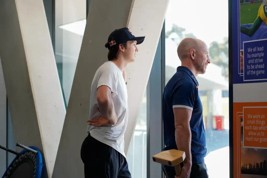 Two people stand looking outside a large window, whilst standing inside the VIS Gym. One person wears a white T-shirt, dark pants, and a black cap. The other person wears a blue collared VIS shirt and holds a small wooden block.
