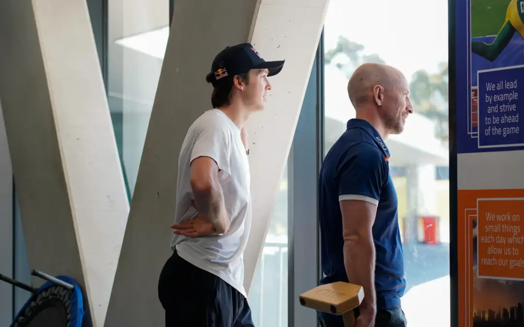 Two people stand looking outside a large window, whilst standing inside the VIS Gym. One person wears a white T-shirt, dark pants, and a black cap. The other person wears a blue collared VIS shirt and holds a small wooden block.
